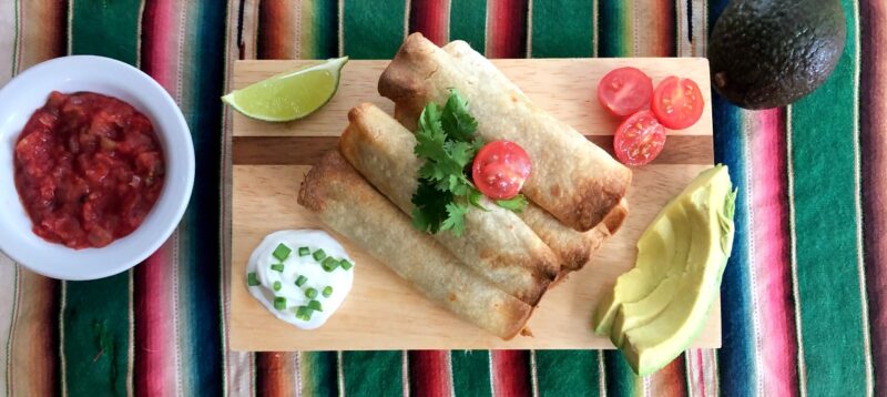 taquitos on a plate with salsa, avocado, sour cream and tomatoes.