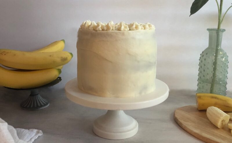 banana cake on a cake stand with bananas and cream cheese frosting