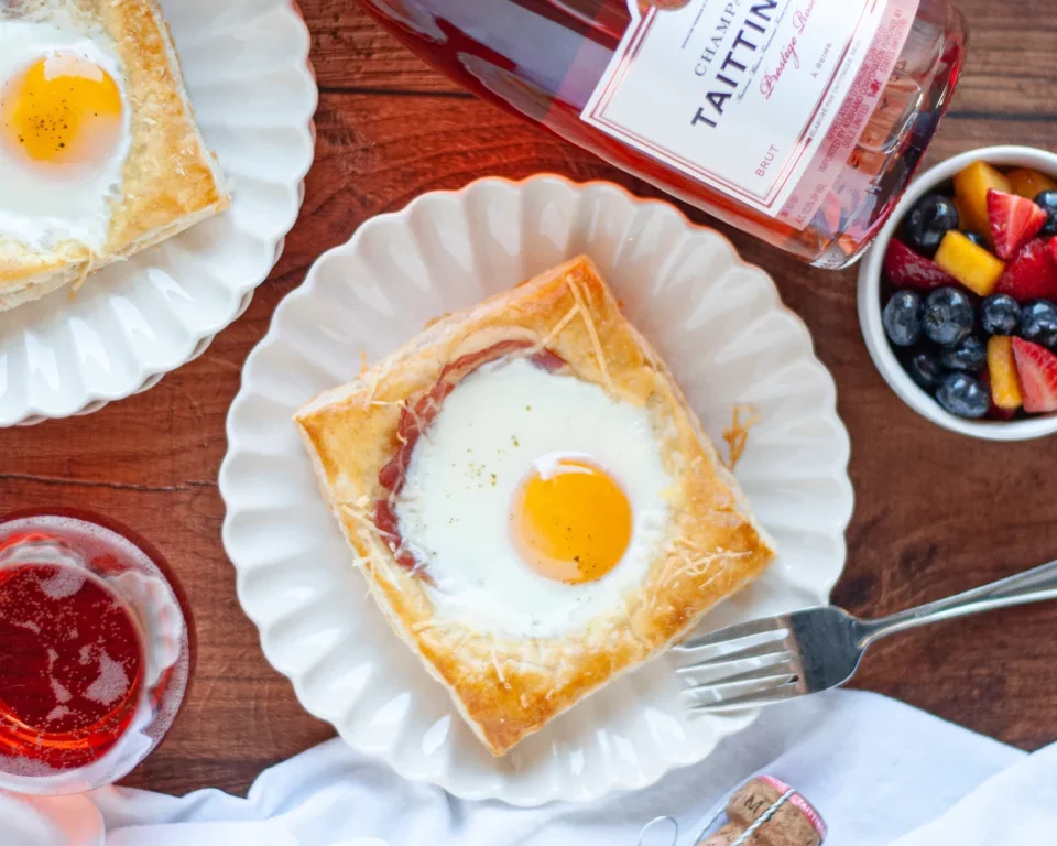 Eggs Benedict served on a white plate with a runny yolk, alongside berries and rosé wine.